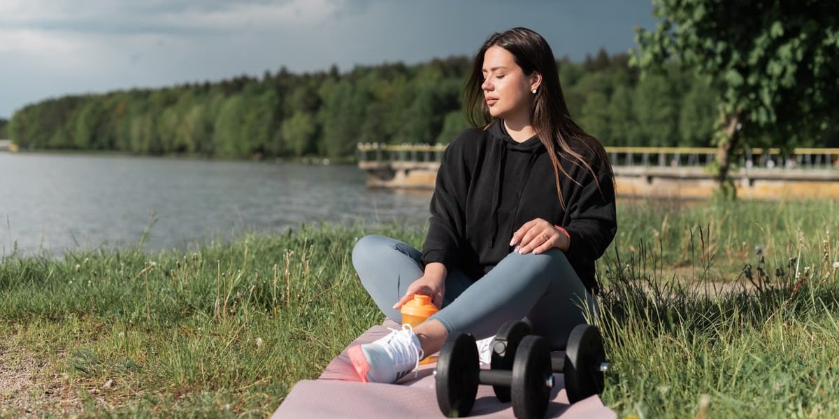 Woman sitting on yoga mat beside a lake with exercise equipment, enjoying a calm outdoor workout.