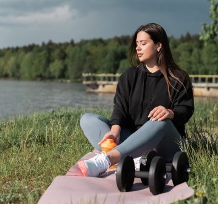 Woman sitting on yoga mat beside a lake with exercise equipment, enjoying a calm outdoor workout.
