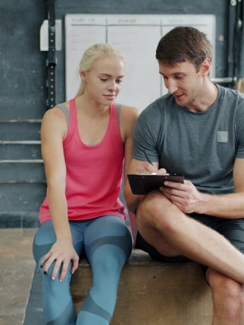 Cheerful sports people man and woman are talking and writing during training break in gym, trainer is planning workout program for female student.