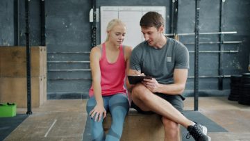 Cheerful sports people man and woman are talking and writing during training break in gym, trainer is planning workout program for female student.