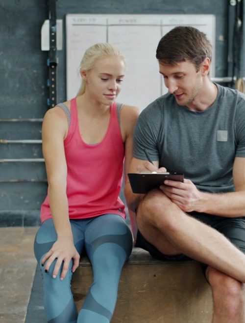Cheerful sports people man and woman are talking and writing during training break in gym, trainer is planning workout program for female student.